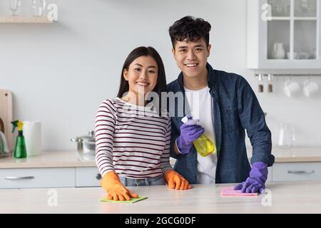 Cheerful millennial asian couple wearing colorful rubber gloves, holding cleaning sprays and dust clothes, smiling at camera while cleaning kitchen ta Stock Photo