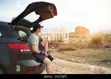 asian photographer sitting at the edge of trunk of a car looking at a deserted castle with camera in hand Stock Photo