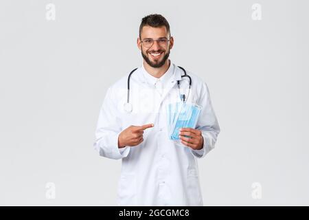 Healthcare workers, medical insurance, pandemic and covid-19 concept. Cheerful, smiling handsome doctor recommend using medical masks, physician in Stock Photo