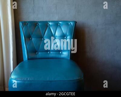 Blue vintage leather sofa seat with pins and buttons decoration near white curtain on grey wall background with copy space. Stock Photo