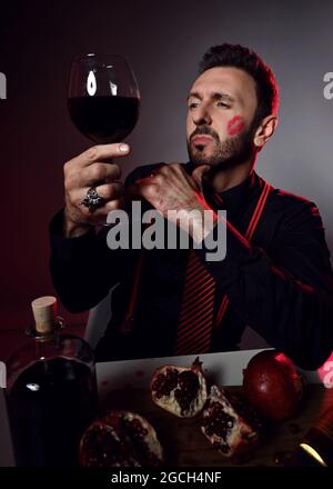 Brutal man in black shirt and trace of kiss on cheek sits at table with pomegranate and juice and loks at glass in hand Stock Photo