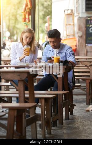 Young woman drinking refreshing cocktail at outdoor cafe and looking at boyfriend texting friends or using app on smartphone Stock Photo