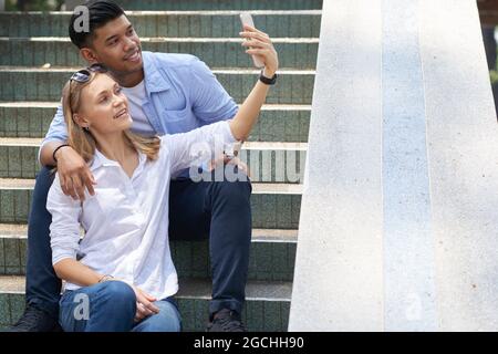 Happy diverse young couple sitting on steps and taking selfie to post on social media Stock Photo
