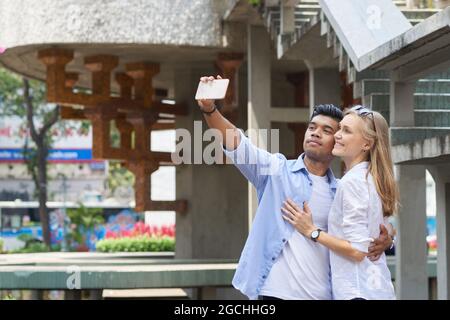 Happy multi-ethnic couple in love hugging and taking selfie on smartphone Stock Photo