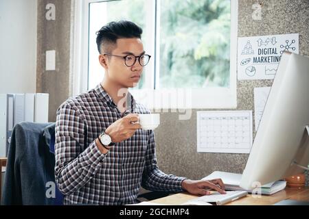 Serious businessman in glasses drinking morning coffee and reading e-mails from colleagues and clients on computer screen Stock Photo