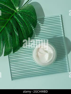 Cosmetic cream or moustirizer on transparent ribbed acrylic plate with tropical palm monstera leaf over blue background. Open round jar, aesthetic swirls cream. Top view flatlay. Copy space. Vertical