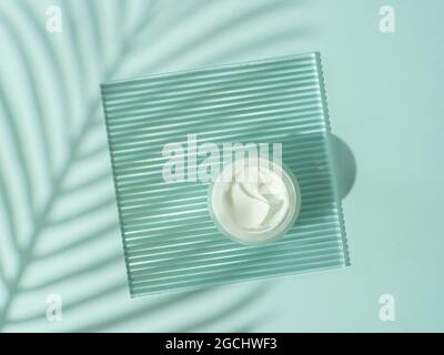 Cosmetic cream or moustirizer on transparent ribbed acrylic plate with tropical palm leaf shadows on blue background. Open round glass jar with aesthetic swirls cream. Top view flatlay. Copy space Stock Photo