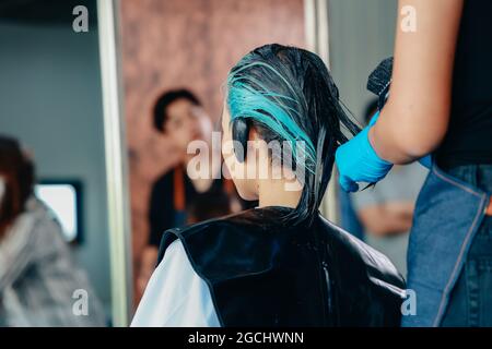 Portrait of young Asian female sitting in modern parlour while getting hair colouring done wearing face mask for protection with female hairdresser colouring hair Stock Photo