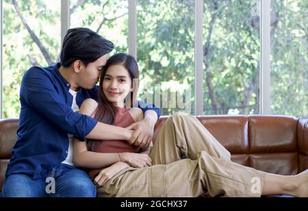 Asian man cuddle his girlfriend with both arms. Young lovers spend time together on holidays in the living room. Stock Photo