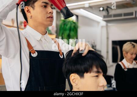 Young male Asian hairdresser wearing apron drying handsome young man's hair in a modern barber shop while concentrating on work Stock Photo