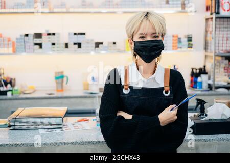 Asian female hairdresser wearing uniform and face mask on in barbershop during Coronavirus pandemic Stock Photo