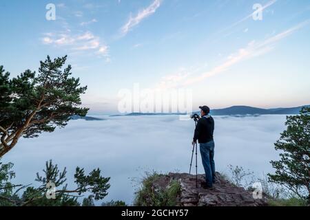 a man photographer in the mountains takes pictures with a camera on a tripod of nature and the morning fog of the hills. the early time is dawn and mi Stock Photo