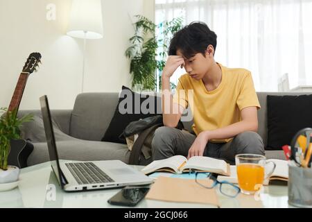 Bored schoolboy tired of doing math homework for school, he is sitting on sofa and looking at laptop screen Stock Photo