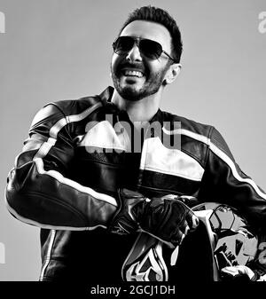 Black and white. Cool bearded man biker in leather jacket and gloves stands holding his motorcycle helmet and laughs Stock Photo