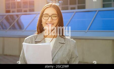 young woman in suit and glasses with papers smiling on the street Stock Photo