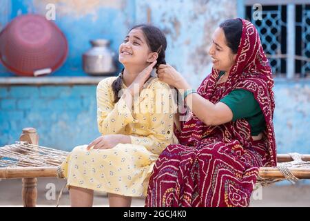 Rural indian mother braids hair of her young adorable daughter, cute girl feel pain as mum getting her ready sitting on traditional bed at village hom Stock Photo