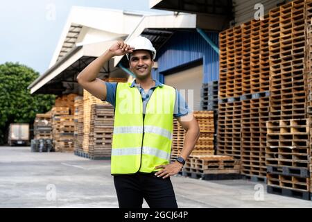 Portrait of young Indian worker working in logistic industry outdoor in front of factory warehouse. Smiling happy man with industrial hard hat looking at camera hands on hip at depot Stock Photo