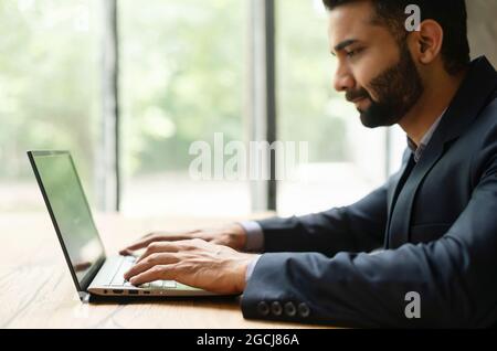 Serious confident young indian man entrepreneur in formal suit working on a laptop sitting at the desk in office, side view of smart eastern businessman typing on the keyboard, checking emails Stock Photo