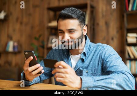 Young handsome Indian freelancer man using smartphone for online payment, holding credit card and inputs data, using app for ordering food, shopping and purchasing online Stock Photo