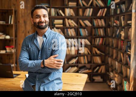 Smiling bearded indian businessman stands near desk and looks at the camera. Young positive male student in library with bookshelves on background. Proud and successful mixed-race small business owner Stock Photo