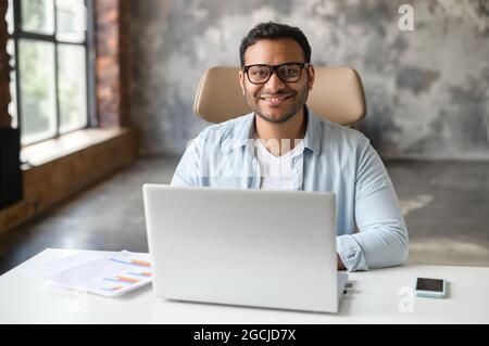 Cheerful indian male student or office worker is using laptop computer sitting at the desk. Friendly and optimistic eastern dark-haired freelancer guy in eyeglasses looks at camera and smiles Stock Photo