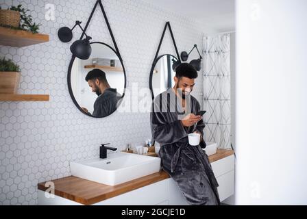 Young man with coffee and bathrobe indoors in bathroom at home, using smartphone. Stock Photo