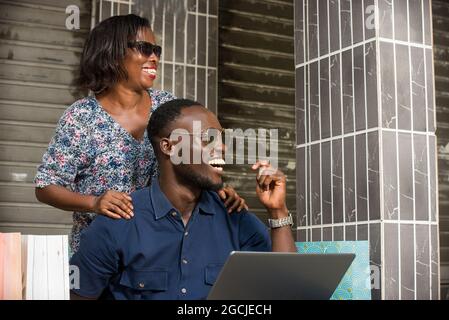 young couple sitting with shopping bags and using a laptop to make online purchases. couple laughing and looking sideways Stock Photo