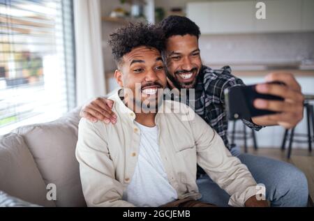 Portrait of young adult brothers on sofa indoors at home, taking selfie. Stock Photo