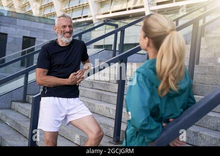 Joyful middle aged guy chatting with sportive woman while standing outdoors after morning workout in the city Stock Photo