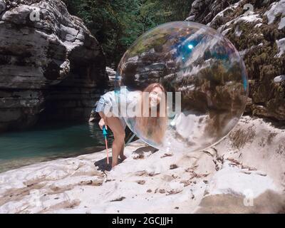 Smiling girl looks through giant soap bubble flying in the canyon gorge near the river Stock Photo