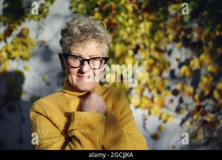 Senior woman standing outdoors against wall in town, looking at camera. Stock Photo