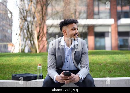 Portrait of young man student sitting outdoors in city, using smartphone. Stock Photo