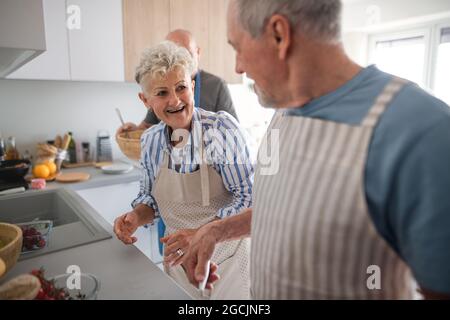 Senior couple with friends having party indoors, talking when cooking. Stock Photo