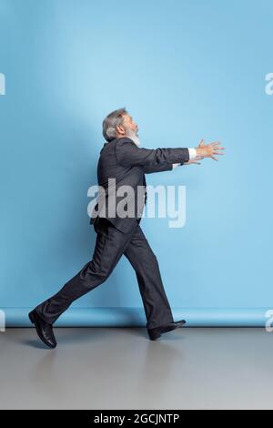 Scared professor, teacher, gray-headed bearded man running, hurrying to class isolated on blue studio background. Funny meme emotions Stock Photo