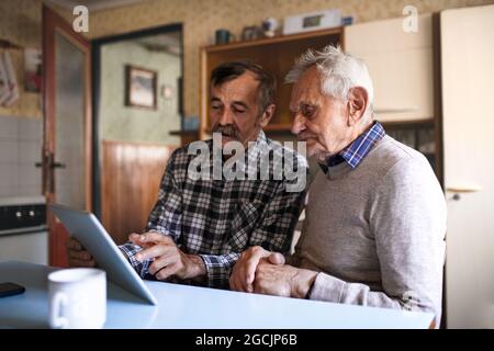 Portrait of man with elderly father sitting at the table indoors at home, using tablet. Stock Photo