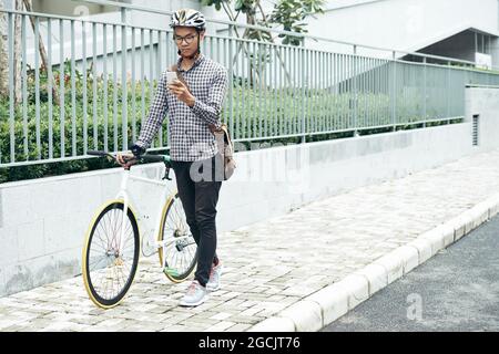Young man with bicycle walking down the street and reading text messages or notifications on his smartphone Stock Photo