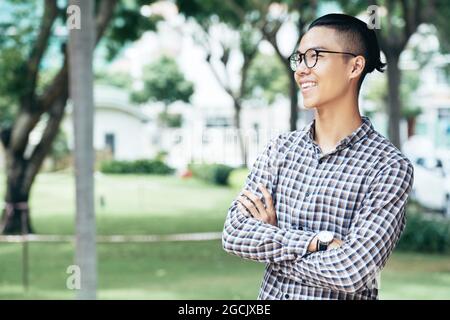 Positive confident young man in plaid shirt standing in city park and looking away Stock Photo