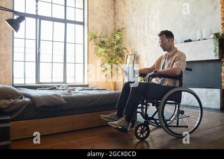 Full length portrait of modern young man in wheelchair using laptop at home in designer interior, copy space Stock Photo