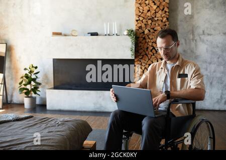 Portrait of modern adult man in wheelchair using laptop at home in designer interior, copy space Stock Photo