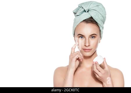Beautiful young woman applying moisturiser face cream, looking at camera smiling. Studio shot on white background. Beauty and skincare concept. Stock Photo