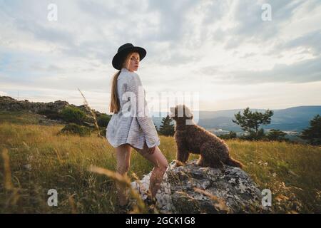 Side view of female owner with obedient Labradoodle dog sitting on rock in mountains looking at camera Stock Photo