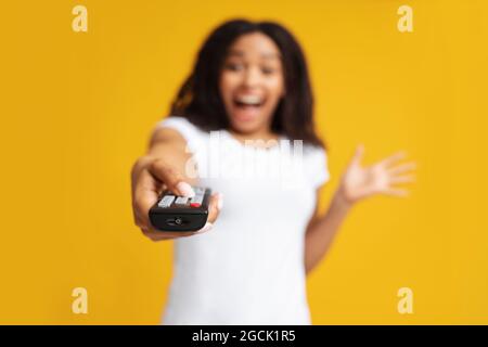 Excited black woman holding remote control in hand, pointing it at camera, watching tv, standing over yellow background Stock Photo