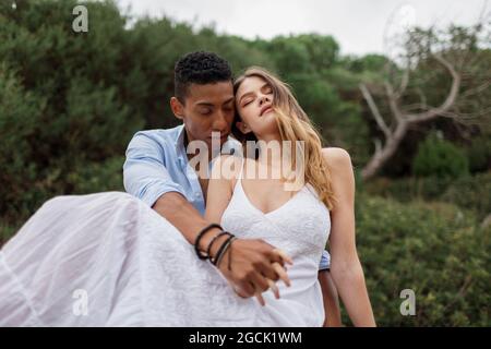 Multiracial couple of newlyweds sitting on stump of tree in woods and hugging on wedding day Stock Photo