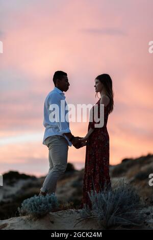 Loving multiracial couple in elegant clothes embracing on hill on background of sundown sky over sea in summer Stock Photo