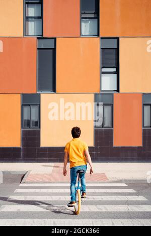 back view of unrecognizable talented man in casual wear sitting on unicycle crossing road on zebra on modern urban street with colorful building