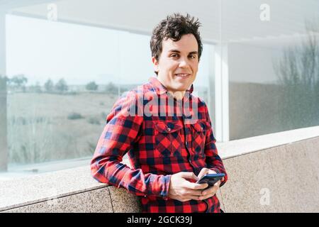 Smiling young male in checkered shirt looking at camera while browsing mobile phone near modern building with glass walls and stone fence Stock Photo