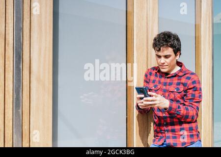 Young male in casual wear using smartphone while standing against modern urban building Stock Photo
