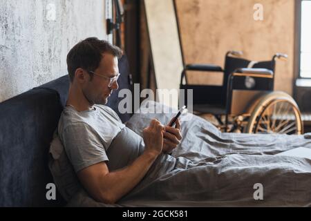 Side view portrait of modern man with disability using smartphone in bed, wheelchair in background, copy space Stock Photo