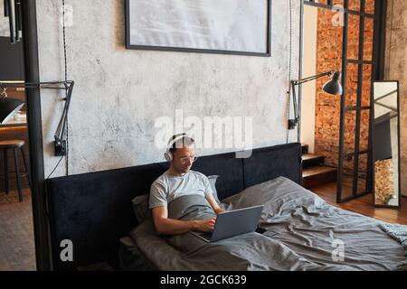 Portrait of modern adult man using laptop in bed and wearing headphones, copy space Stock Photo