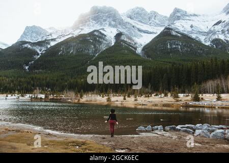 Back view of unrecognizable female hiker standing alone on shore of Quarry Lake against majestic forested mountains with snow covered rocky peaks in B Stock Photo